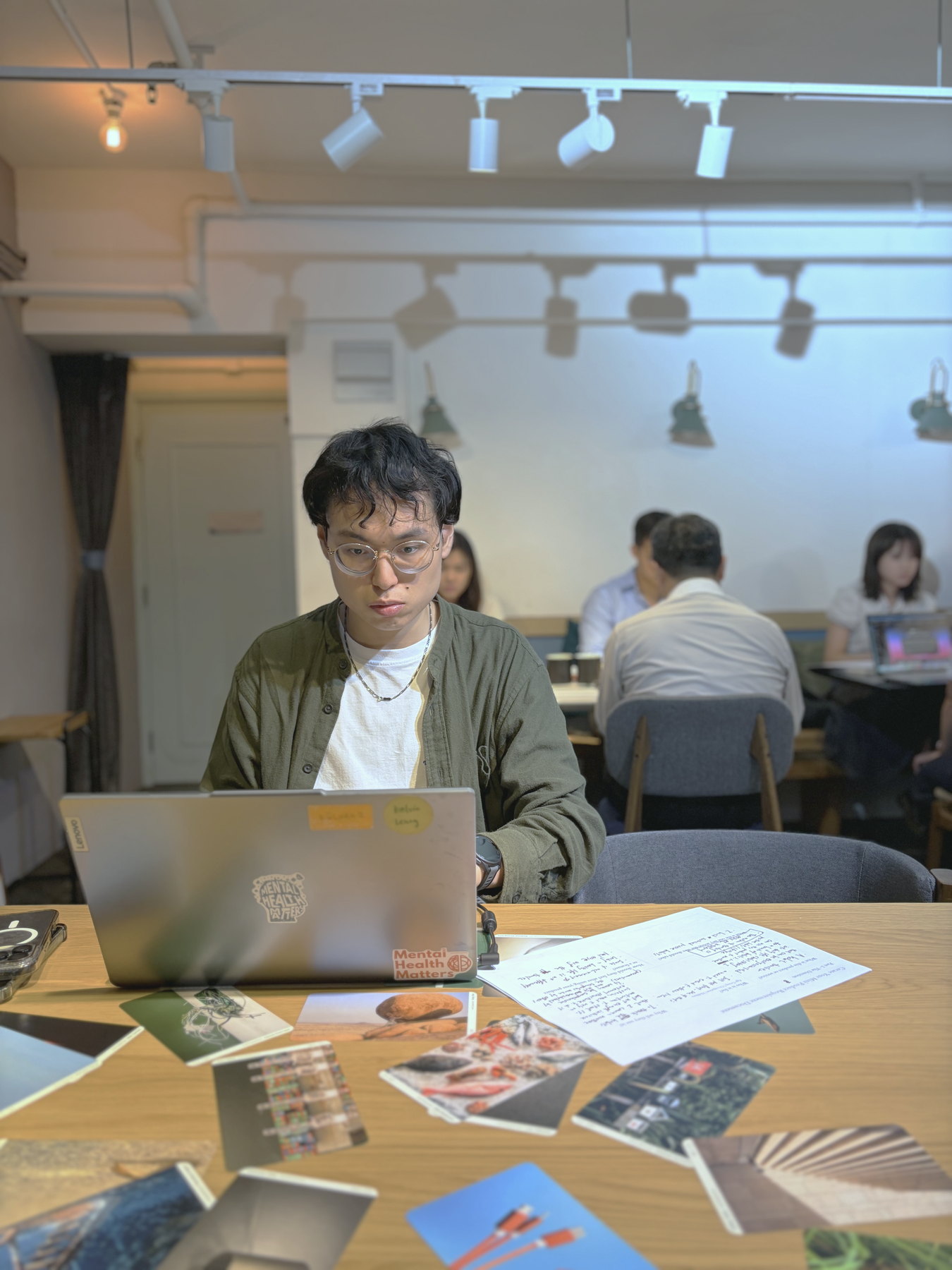 A participant working intently at a laptop, printed image cards spread across the table in front of him and others building behind.