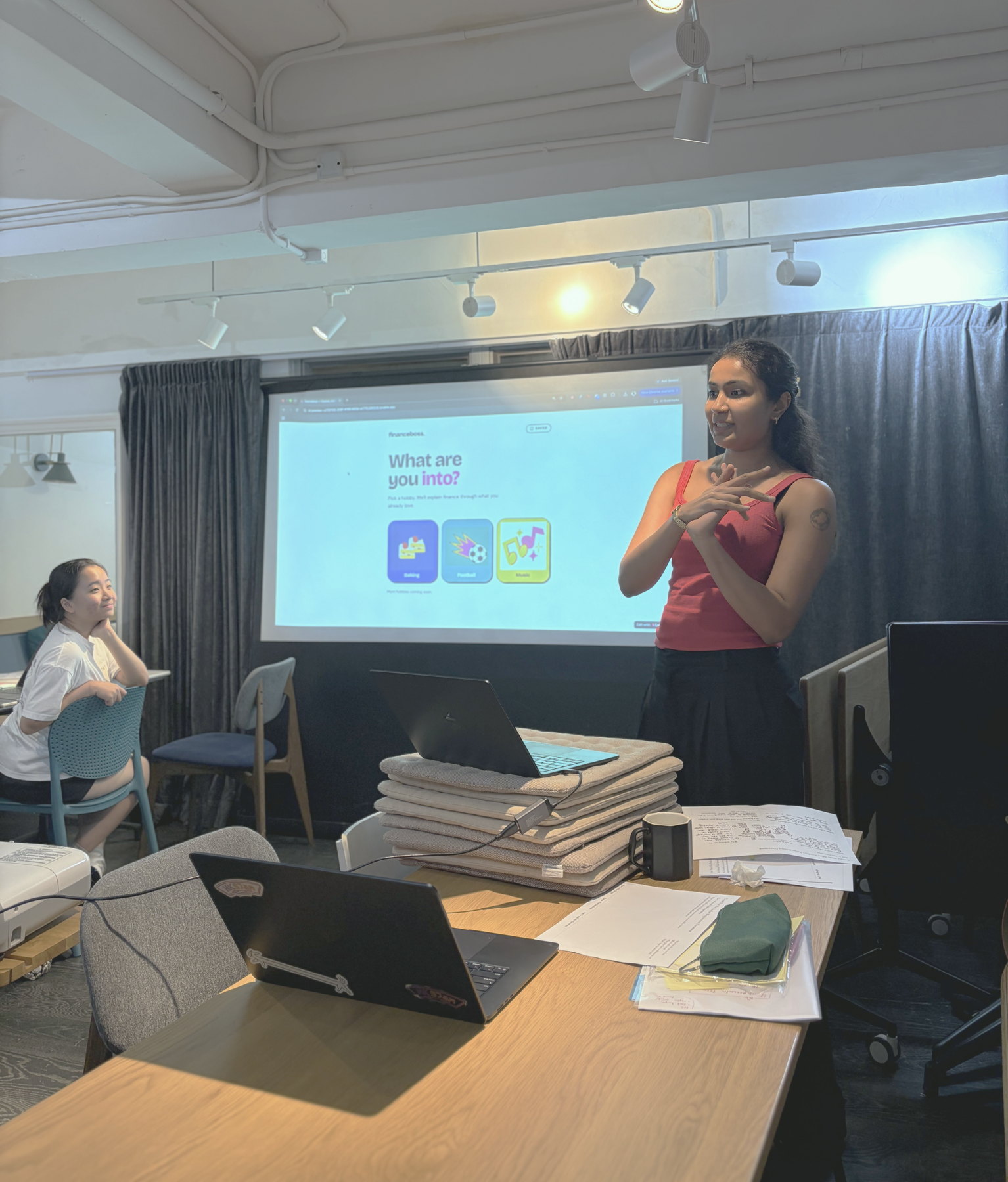A participant standing beside a projected screen showing the app she built, presenting it to the room while another participant watches from a nearby seat.