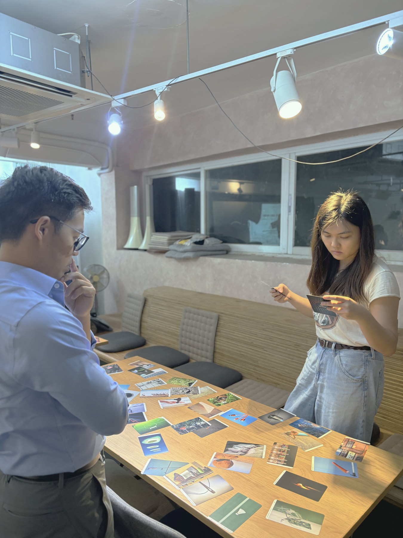 Two participants standing over a table of printed image cards, one holding a card and considering it, the other with a hand to his chin.