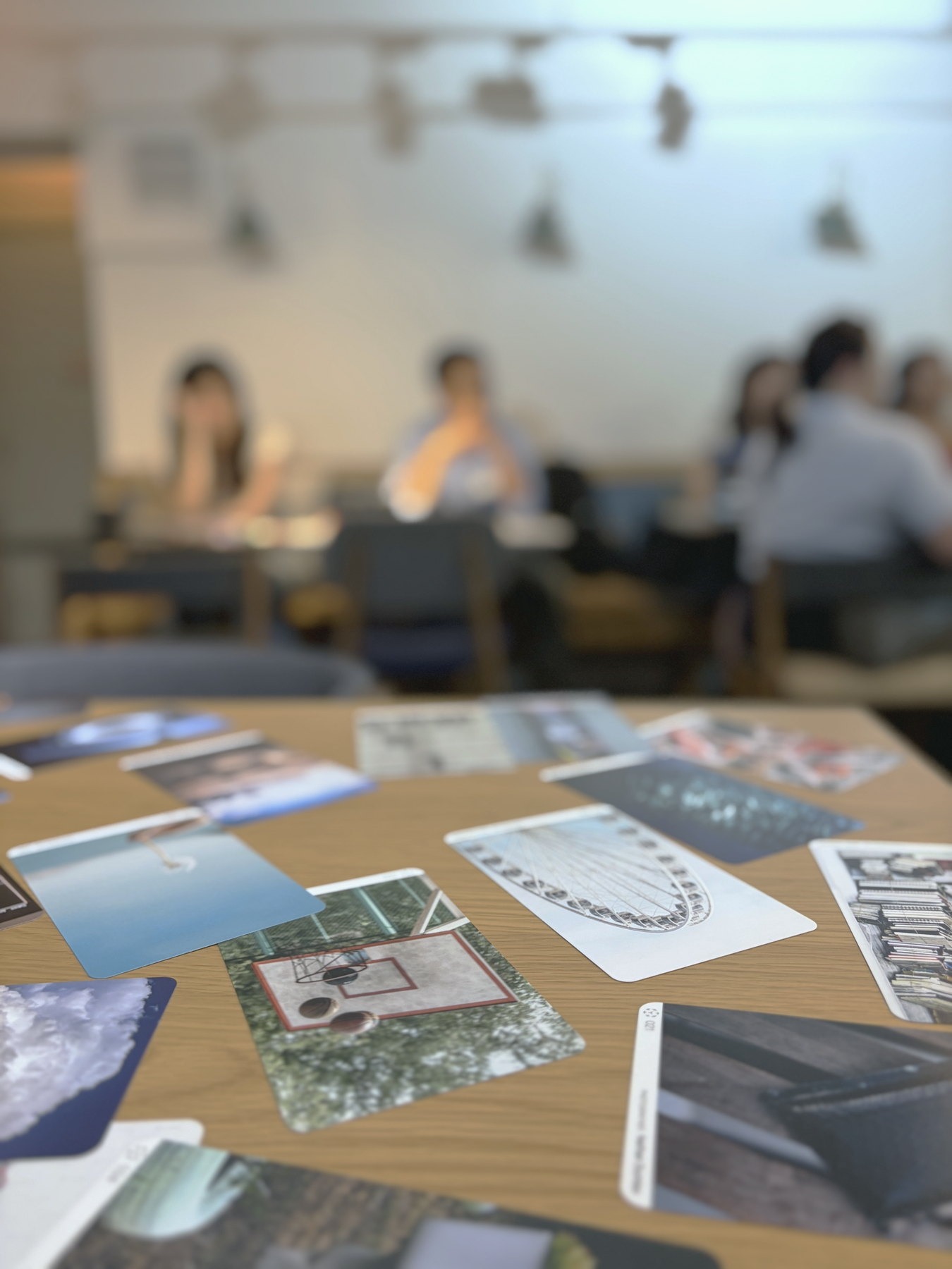 Printed image cards spread across a wooden table in sharp focus, the workshop and its participants soft and out of focus behind.