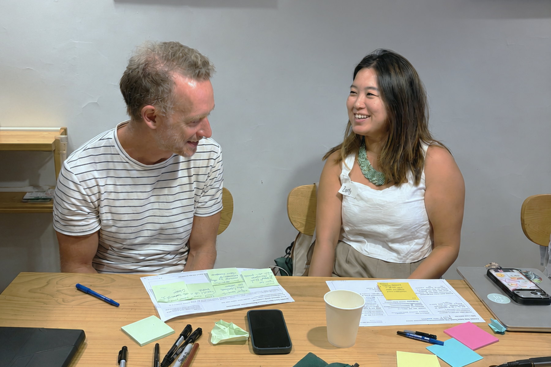 Two workshop participants laughing mid-conversation across a wooden table covered in handwritten worksheets. No laptops are open.